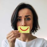 Woman Hiding Pain, Frowning with a picture of a smile in front of her face