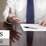IRS official gesturing v with documents and pen