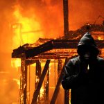 Hooded figure standing before a burning building