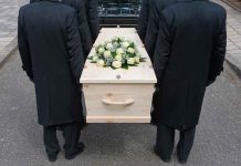 Four people carrying a coffin with white flowers