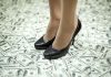 A pair of black high heels standing on a floor covered with dollar bills