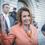 Woman in orange blazer speaking surrounded by security personnel