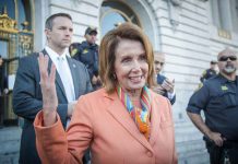 Woman in orange blazer speaking surrounded by security personnel