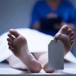 A deceased body on a table with a tag on the foot and a medical professional in the background