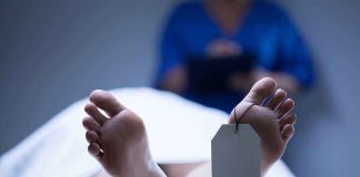 A deceased body on a table with a tag on the foot and a medical professional in the background
