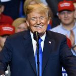 Man in suit speaking at rally with red hats.