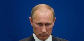 A serious-looking man in a suit at a press conference with a blue background