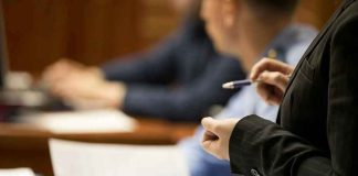 Person holding pen, speaking to seated individuals with papers.