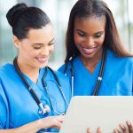 Two nurses in blue scrubs smiling while looking at a tablet