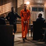 Defendant in orange jumpsuit being escorted by a police officer in a courtroom