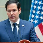 Rubio’s Munich Blast Sparks Furious Backlash A man in a blue suit speaking into a microphone with an American flag in the background
