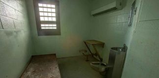 Empty jail cell with bed, barred window, and stainless steel toilet