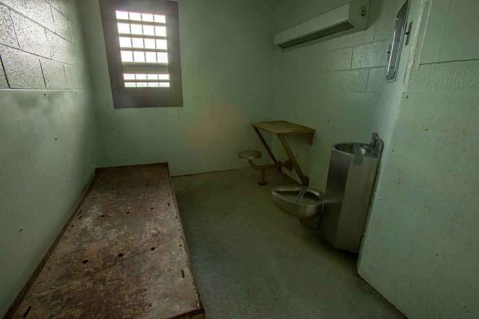 Empty jail cell with bed, barred window, and stainless steel toilet