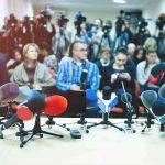 What Did Bill Gates Trade With Epstein? Row of microphones at a press conference with audience and cameras behind