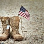 Iran War Opens a Rift With Veterans Combat boots with dog tags and a small American flag on gravel