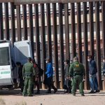 16 Months of Zero Border Releases Border agents escort a group near a tall fence and a patrol van