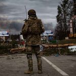 Soldier in camouflage stands at roadblock with smoke in the distance