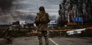 Soldier in camouflage stands at roadblock with smoke in the distance
