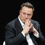 Musk Says MONEY Could Become Almost Meaningless Elon Musk seated in a dark setting with hands steepled near his face