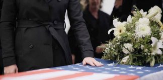 Mourner rests hand on flag-draped casket with flower arrangement