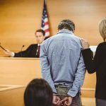 Political Hitman Caught — Death Off Table Defendant in handcuffs stands before a judge in a courtroom