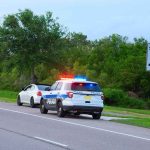 Police SUV with lights on behind a white car on roadside near 45 mph sign