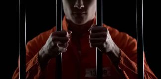 Prisoner in orange jumpsuit gripping jail bars