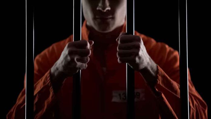 shutterstock_1341238571 Prisoner in orange jumpsuit gripping jail bars