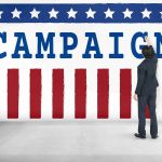 Vanished Democrat’s FLU Excuse Questioned After New Audio Man in suit writing on large campaign banner with stars and stripes